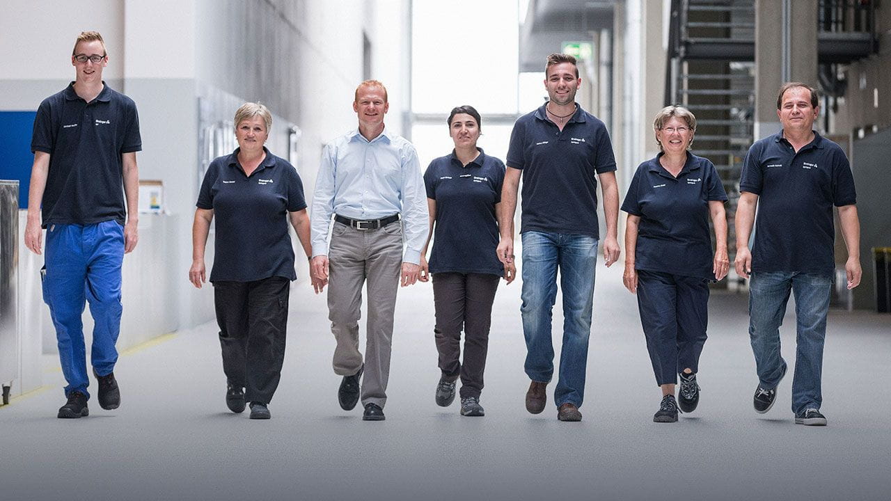7 Ensinger Mitarbeiter laufen in einer Werkhalle in einer Reihe auf Betrachter zu